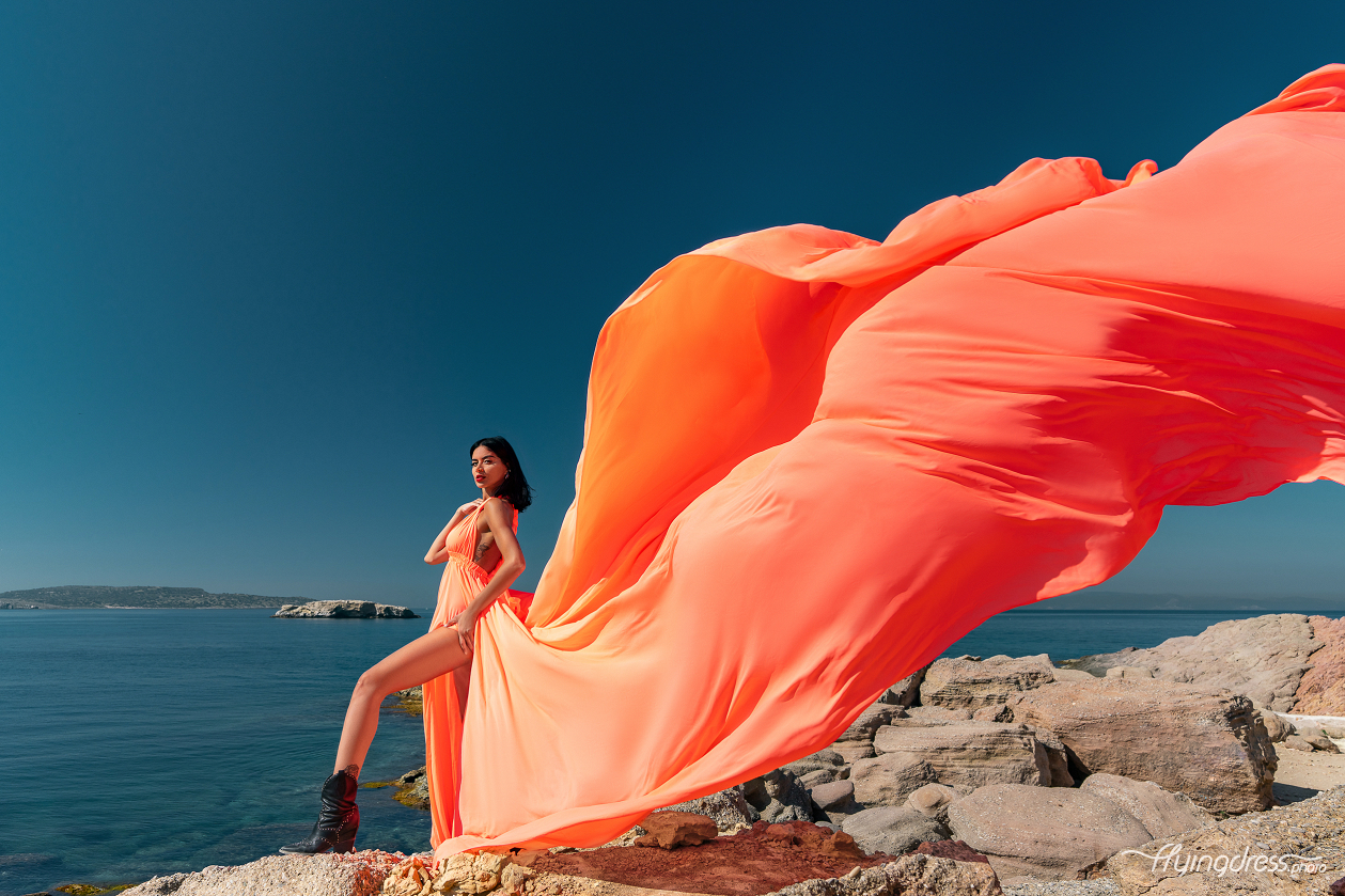 A woman in a flowing orange dress strikes a dynamic pose on the rocky coastline of Vouliagmeni, Athens, with the vibrant fabric of her dress sweeping dramatically in the wind against the backdrop of calm blue waters and a clear sky.